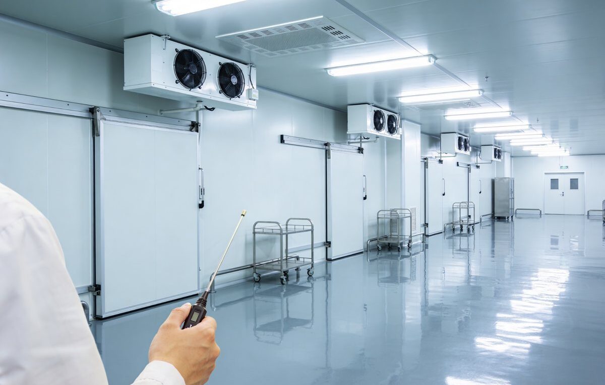 Técnico verifica temperatura em corredor com portas de câmaras frias, evaporadores e equipamentos de refrigeração.