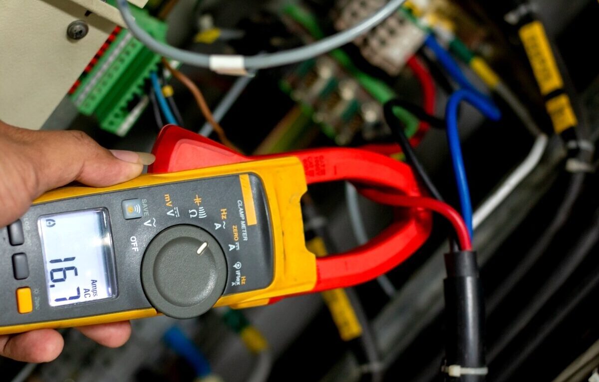 Mão segura pinça amperimétrica ao redor de um fio em painel elétrico durante diagnóstico de ar-condicionado