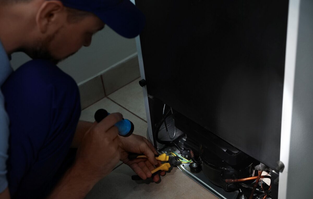 Técnico inspeciona relé, protetor térmico e compressor na parte traseira de uma geladeira que liga e desliga rápido.